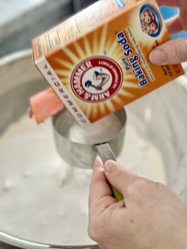 A person pouring baking soda from a box into a measuring cup filled with flour.