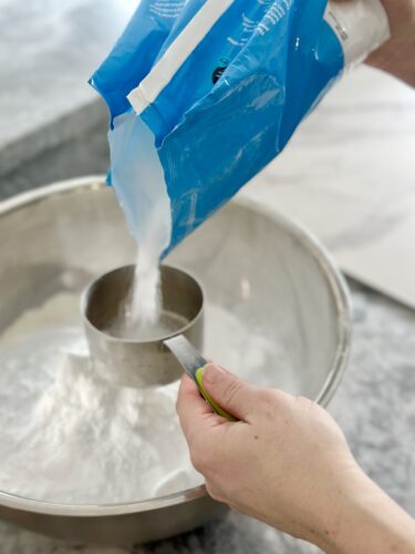 A person's hand pouring white powder from a blue bag into a metal measuring cup over a mixing bowl.