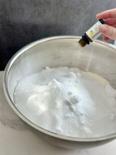 A hand holding a small essential oil bottle over a bowl filled with white powder, possibly for a DIY project.