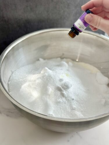 A hand pouring essential oil into a large bowl filled with white powdery ingredients, possibly for a DIY project or recipe.