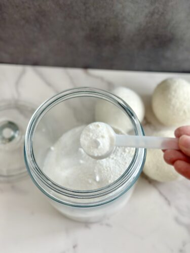 A hand holding a white measuring spoon full of powder above a glass jar filled with the same powder, with white round bath bombs in the background.