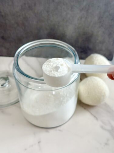 A clear glass jar containing white powder, with a measuring spoon filled with the powder in the foreground and textile balls placed beside the jar.