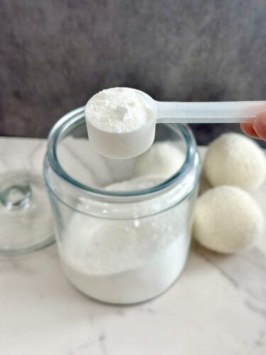 A hand holding a measuring scoop filled with white powder above a glass jar of the same powder, with white spheres in the background.