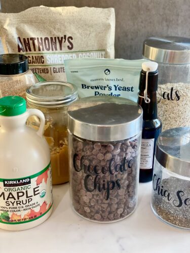 A collection of various cooking and baking ingredients including organic shredded coconut, chocolate chips, oats, chia seeds, maple syrup, peanut butter, vanilla extract, and brewer's yeast powder, arranged on a countertop.