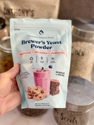 A person holding a bag of Brewer's Yeast Powder labeled 'mommy knows best,' showcasing a pink smoothie and cookies on the package.
