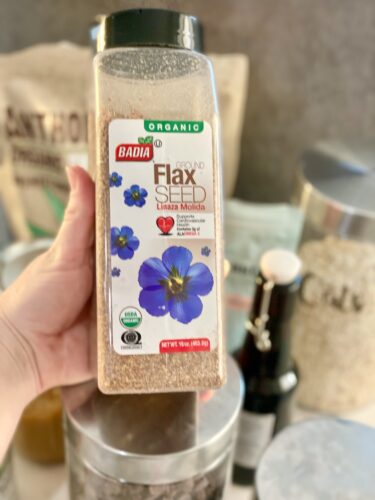 Bottle of organic ground flax seed held in a hand, featuring a label with blue flowers and nutritional information.