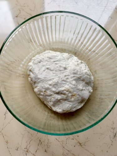 A round ball of dough resting in a clear glass bowl, on a light-colored surface.