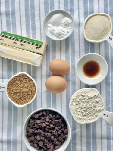 Ingredients for baking: chocolate chips, brown sugar, granulated sugar, baking soda, eggs, vanilla extract, flour, and butter arranged on a striped cloth.