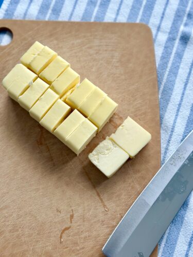 Cubes of yellow butter on a wooden cutting board with a knife beside them, against a blue and white striped background.