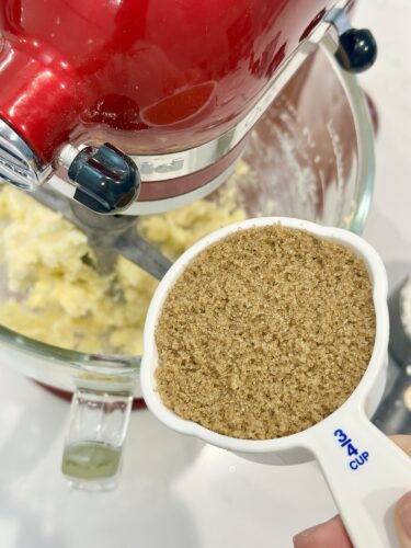 Close-up of a measuring cup with brown sugar, held above a mixing bowl with creamed butter and sugar, featuring a red stand mixer in the background.