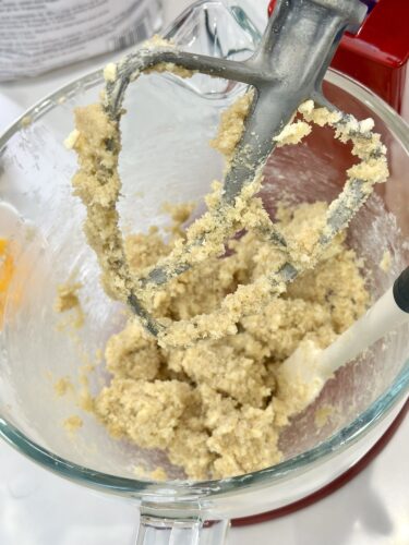 A mixing bowl with cookie dough being blended by an electric mixer, showing a paddle attachment coated with the mixture.