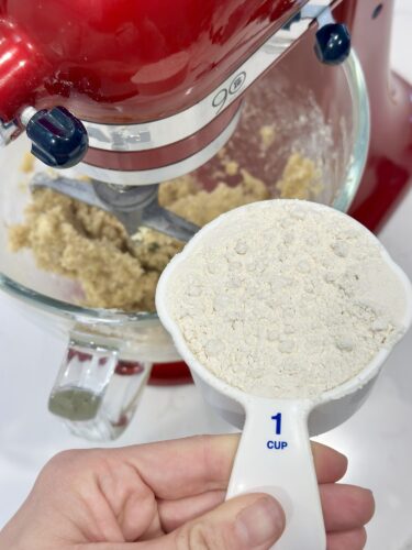 A hand holding a measuring cup filled with flour, with a red stand mixer and cookie dough mixture in the background.