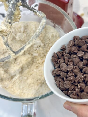 A bowl of chocolate chips held above a mixing bowl containing cookie dough batter, with a mixer attachment in the background.