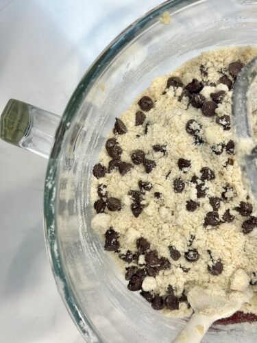 Close-up view of a glass mixing bowl containing cookie dough with chocolate chips and a spatula, showing a crumbly texture.