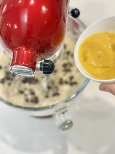A red stand mixer is in the foreground, mixing ingredients in a glass bowl, while a hand holds a small white bowl containing beaten eggs.