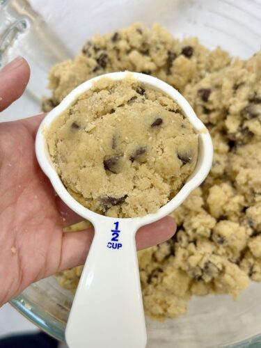 A hand holding a half-cup measuring spoon filled with chocolate chip cookie dough, with more dough in the background.