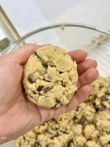 A person's hand holding a round ball of cookie dough with chocolate chips, above a bowl of cookie dough.