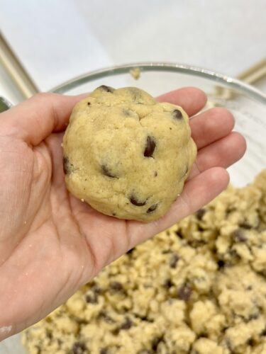 A person's hand holding a cookie dough ball, with more cookie dough scattered in a bowl in the background.