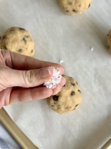 A hand holding a pinch of coarse salt above cookie dough balls on a baking sheet.