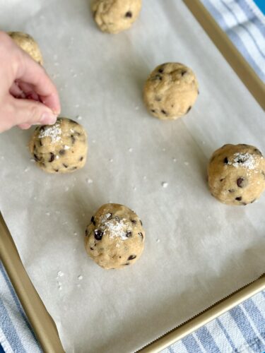 A hand sprinkling powdered sugar on cookie dough balls on a parchment-lined baking sheet.