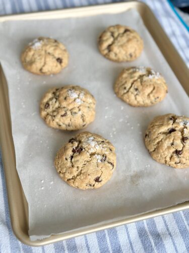 Freshly baked cookies on a parchment-lined baking tray, featuring six round cookies with chocolate chips and a sprinkle of sea salt.
