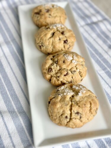 A platter with four freshly baked chocolate chip cookies, lightly dusted with powdered sugar, on a striped cloth background.