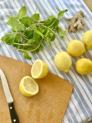 Sliced lemon on a cutting board, surrounded by whole lemons, fresh mint leaves, and ginger root.
