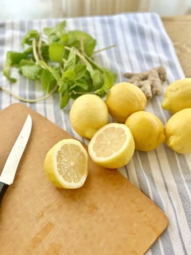 A wooden cutting board with a knife, fresh lemons, a piece of ginger, and green mint leaves on a striped tablecloth.