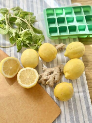 A collection of fresh lemons, including whole and halved lemons, arranged on a surface with ginger root and mint leaves, alongside a green ice cube tray.