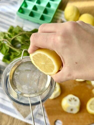 A hand squeezing a lemon over a fine mesh strainer, with lemon slices and mint leaves in the background.
