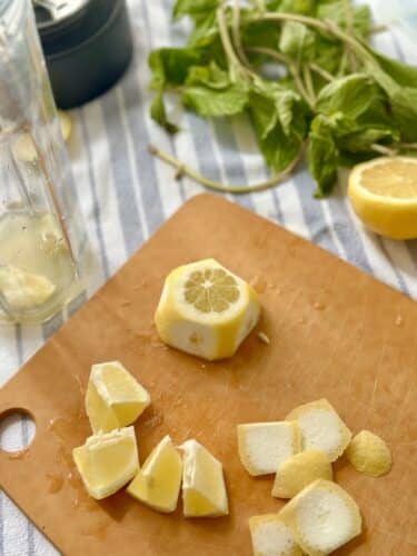 A cutting board with chopped lemon pieces and a whole lemon next to fresh mint leaves, with a glass blender in the background.