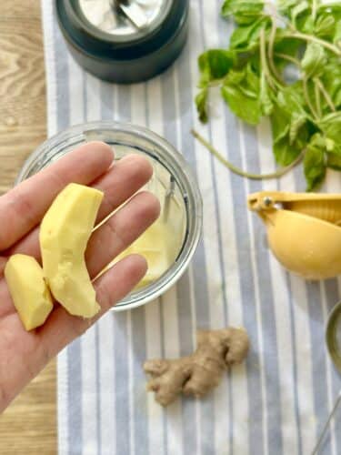 A hand holding pieces of ginger, with a glass jar and fresh herbs in the background.