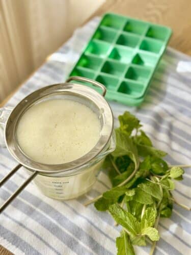 A glass measuring cup with strained liquid, a fine mesh sieve resting on the cup, fresh mint leaves nearby, and a green ice cube tray on a striped cloth.