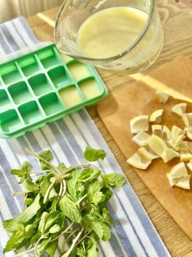 A measuring cup pouring liquid into a green ice cube tray, with fresh mint leaves and chopped bananas on a wooden cutting board beside it. A striped cloth is underneath.