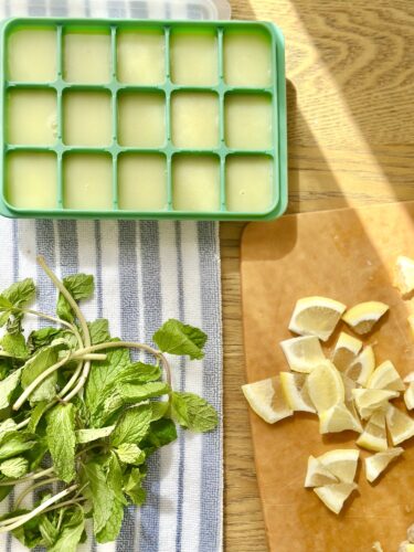 A green ice cube tray filled with lemonade sits on a striped kitchen towel next to a wooden cutting board with lemon pieces and fresh mint leaves.