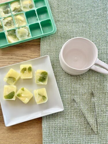 A plate of frozen lime and mint ice cubes on a wooden table, next to an empty pink mug and an ice cube tray.