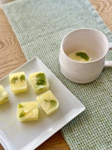 A plate of frozen lemon-lime cubes with mint leaves, next to a white cup containing a drink with a mint leaf, on a textured green napkin.