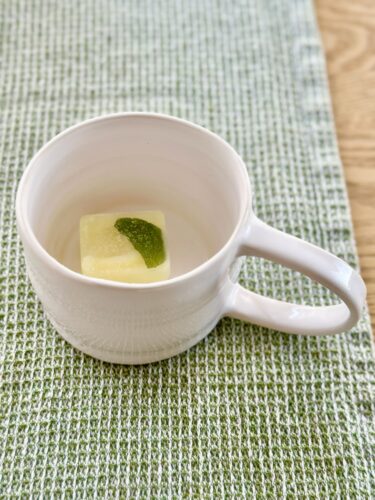 A white ceramic cup with a square piece of yellow food and a small green garnish inside, positioned on a textured green placemat.