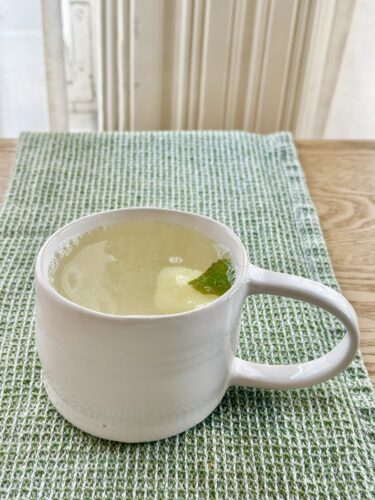 A white ceramic mug filled with a light-colored beverage, featuring a slice of fruit and a mint leaf, placed on a textured green placemat on a wooden table.