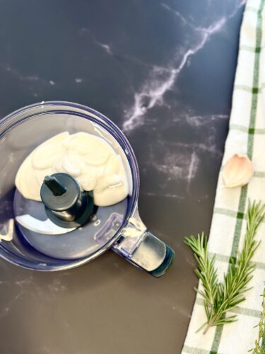 A top view of a food processor containing a creamy mixture, placed on a dark surface. Nearby are fresh garlic, rosemary sprigs, and a checkered dish towel.