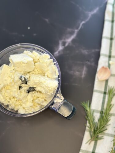 A food processor containing soft cheese with a piece of butter and a sprig of rosemary, placed on a dark countertop next to a green and white checkered cloth and a clove of garlic.