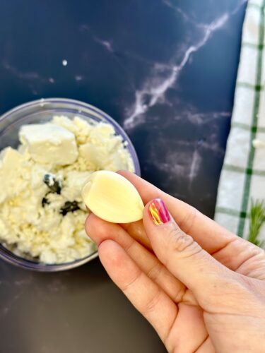 A hand holding a clove of garlic above a bowl of ricotta cheese with herbs.