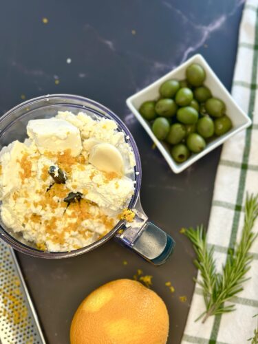 A bowl of crumbled feta cheese topped with orange zest and herbs, accompanied by a small dish of green olives, an orange, and a sprig of rosemary on a dark countertop.