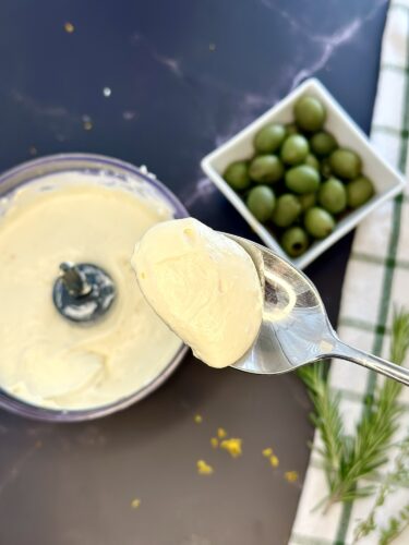 A spoonful of creamy mayonnaise held above a bowl of mayonnaise, with a dish of green olives in the background and a sprig of rosemary on the side.