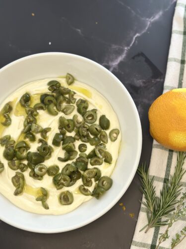 A white bowl filled with a creamy dip topped with sliced green olives, drizzled with olive oil. An orange sits beside the bowl, with a sprig of rosemary and a green checkered napkin.