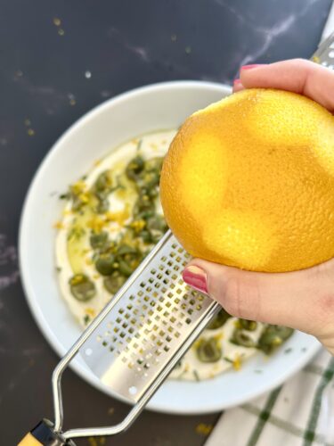 A hand holding an orange over a grater, zesting the fruit into a bowl with cream and green herbs.