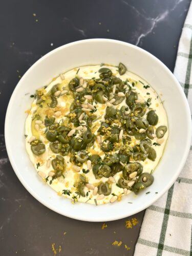 A white bowl filled with a creamy dish topped with green olives, pine nuts, and herbs, presented on a dark surface alongside a checkered cloth.