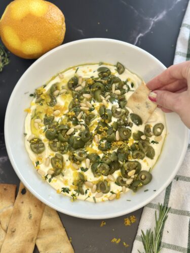 A bowl of creamy dip topped with green olives, pine nuts, and fresh herbs, with an orange on the side and a hand dipping a piece of flatbread into the dip.