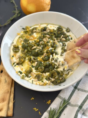 A bowl of creamy yogurt topped with green olives, pine nuts, and herbs, with a hand dipping a piece of flatbread into it. An orange and a sprig of thyme are visible in the background.