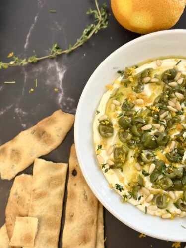 A bowl of hummus topped with green olives, pine nuts, and herbs, accompanied by crispy flatbread and an orange on a dark surface.